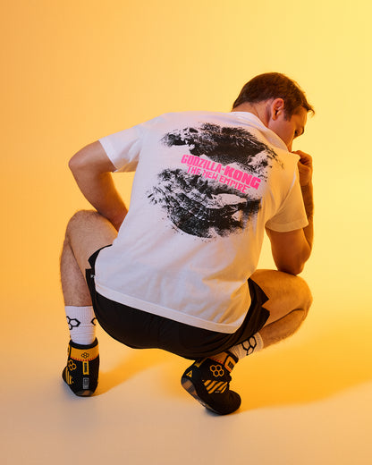 A man crouching from behind wearing a white t-shirt with a large black and pink graphic print on the back, black shorts, and black climbing shoes with yellow details, set against a warm yellow studio background.