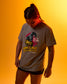 A model wearing a heather gray graphic t-shirt with a 'King of the Mat' wrestling design, posed against a warm orange-yellow gradient background.