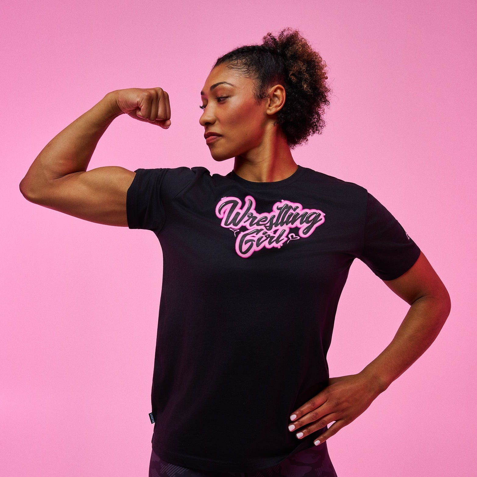 A woman posing in a black short-sleeve t-shirt with 'Wrestling Girl' printed in pink and white script across the chest, paired with dark patterned leggings, set against a solid pink studio background.