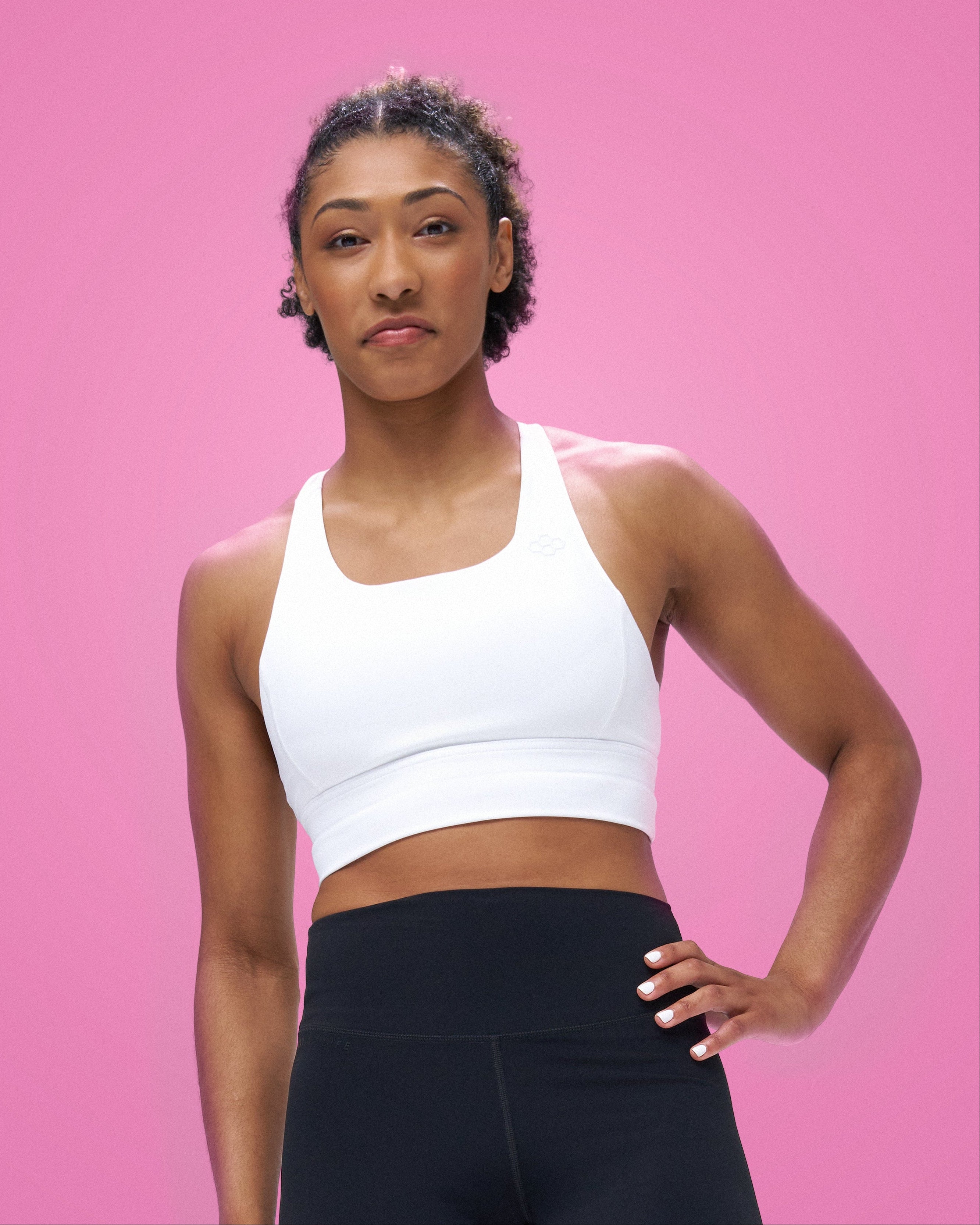 A woman, Kennedy Blades, modeling a RUDIS white scoop-neck athletic sports bra against a solid pink background.