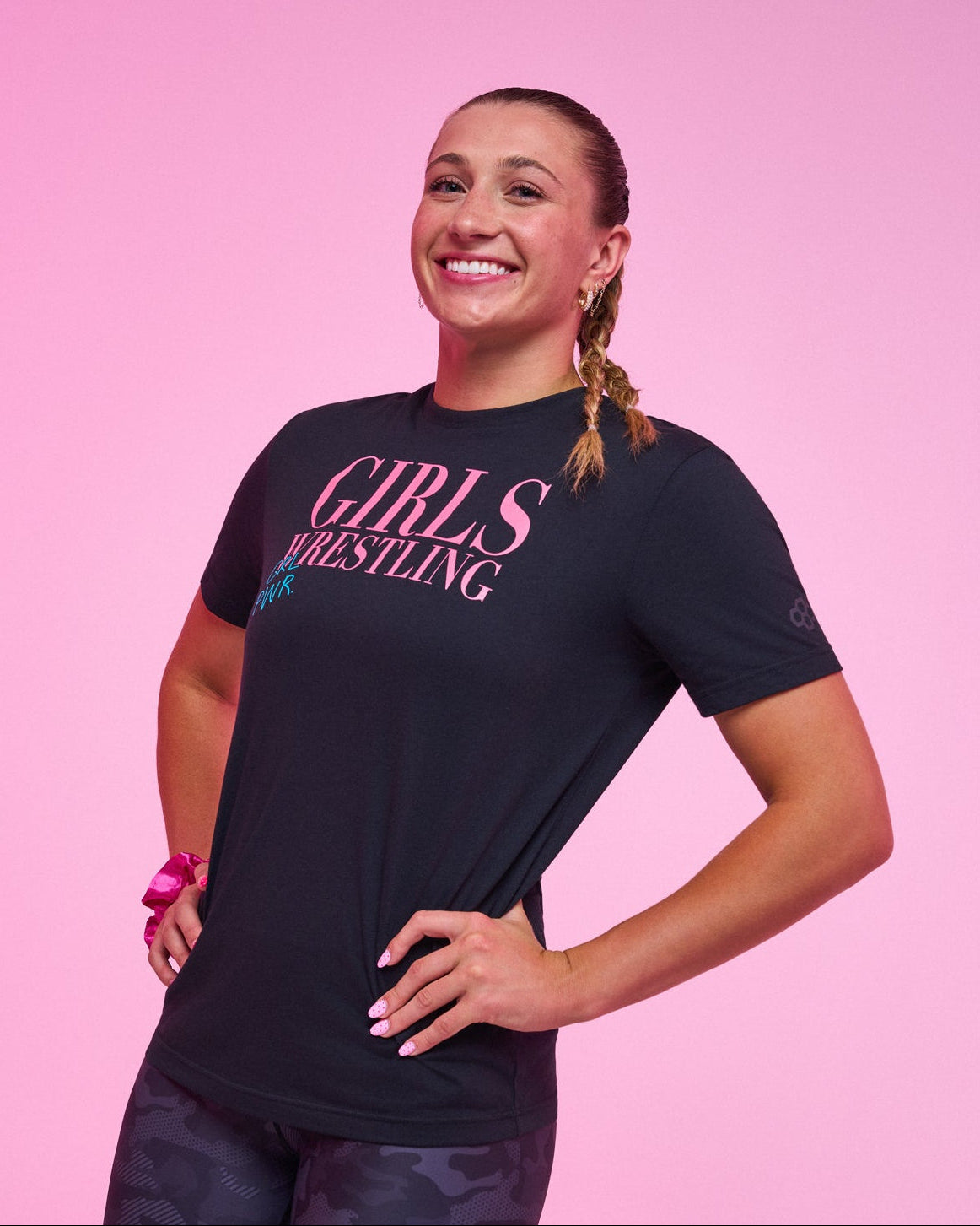 A woman, Kylie Welker, posing in a black RUDIS graphic t-shirt that reads 'GIRLS WRESTLING' in pink, paired with dark camouflage leggings, set against a solid pink background.