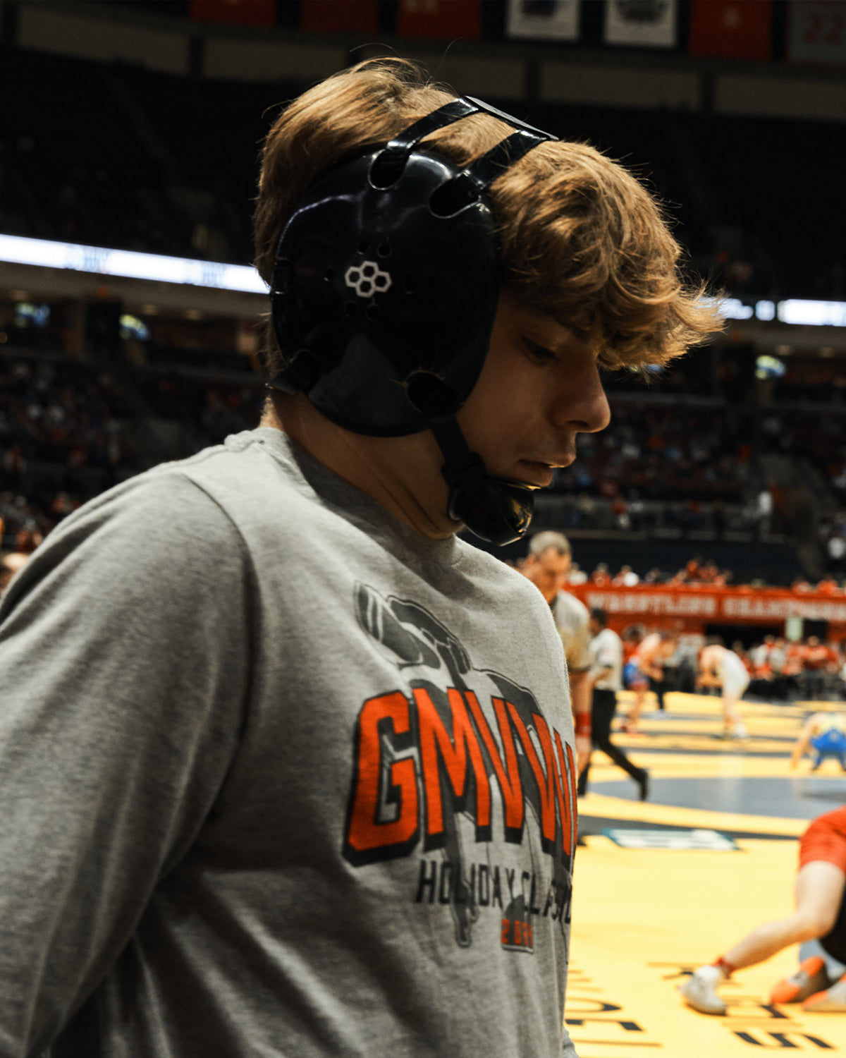 Male wrestler in a GMVWA Holiday Classic t-shirt wearing black RUDIS headgear with wrestlers on the mat in the background. 