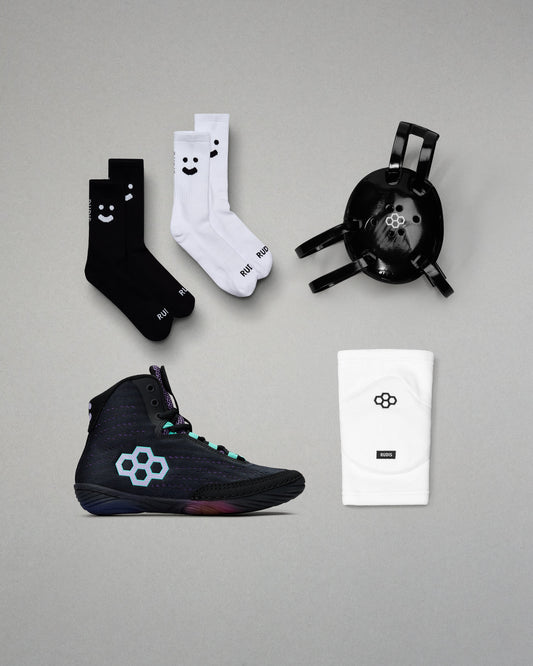 Black wrestling shoes with a white logo, black socks with smiley faces, white socks with smiley faces, and a black headgear on a gray background.