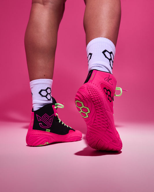 A person's legs wearing bright pink and black athletic wrestling shoes with white crew socks, shown from the calves down against a solid pink studio background.