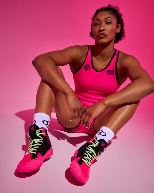 A woman sits cross-legged against a bright pink studio background, wearing a neon pink athletic wrestling singlet and shorts with black trim, black wrestling shoes with bright pink and neon green details, and white socks.