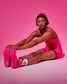 A woman in a bright pink athletic wrestling singlet and matching pink wrestling shoes sits on a pink studio background, reaching toward her foot. She is wearing white socks with black and pink floral patterns.