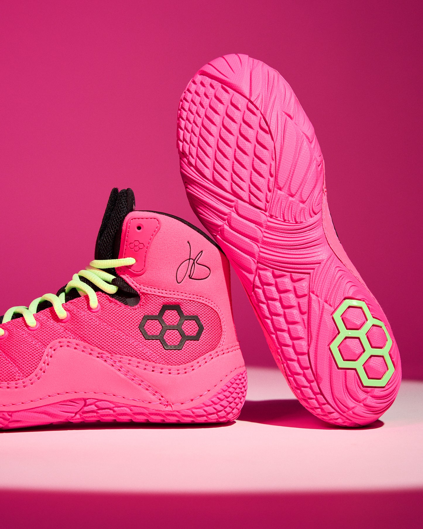 A pair of bright pink athletic wrestling shoes with neon yellow-green laces and a black hexagonal logo, shown on a matching pink studio background. One shoe is positioned upright to display the textured sole, and the other is angled to show the side profile.