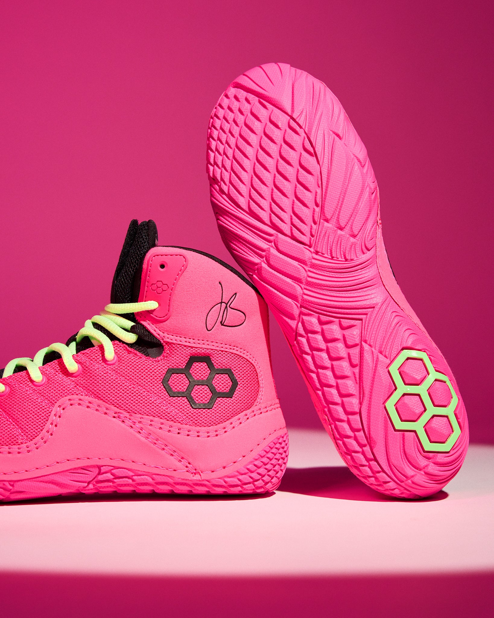 A pair of bright pink athletic wrestling shoes with neon yellow-green laces and a black hexagonal logo, shown on a matching pink studio background. One shoe is positioned upright to display the textured sole, and the other is angled to show the side profile.