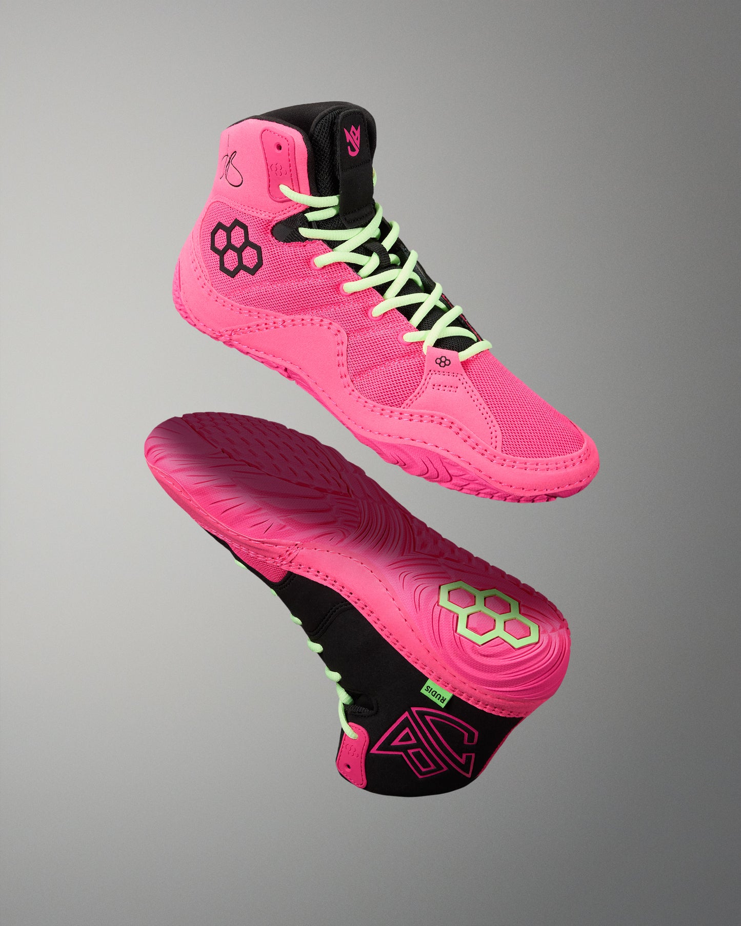 A pair of high-top athletic wrestling shoes in bright pink and black, shown floating against a neutral gray gradient background. One shoe is oriented to show the side profile with neon green laces, while the other shows the sole with a matching pink and black tread pattern.