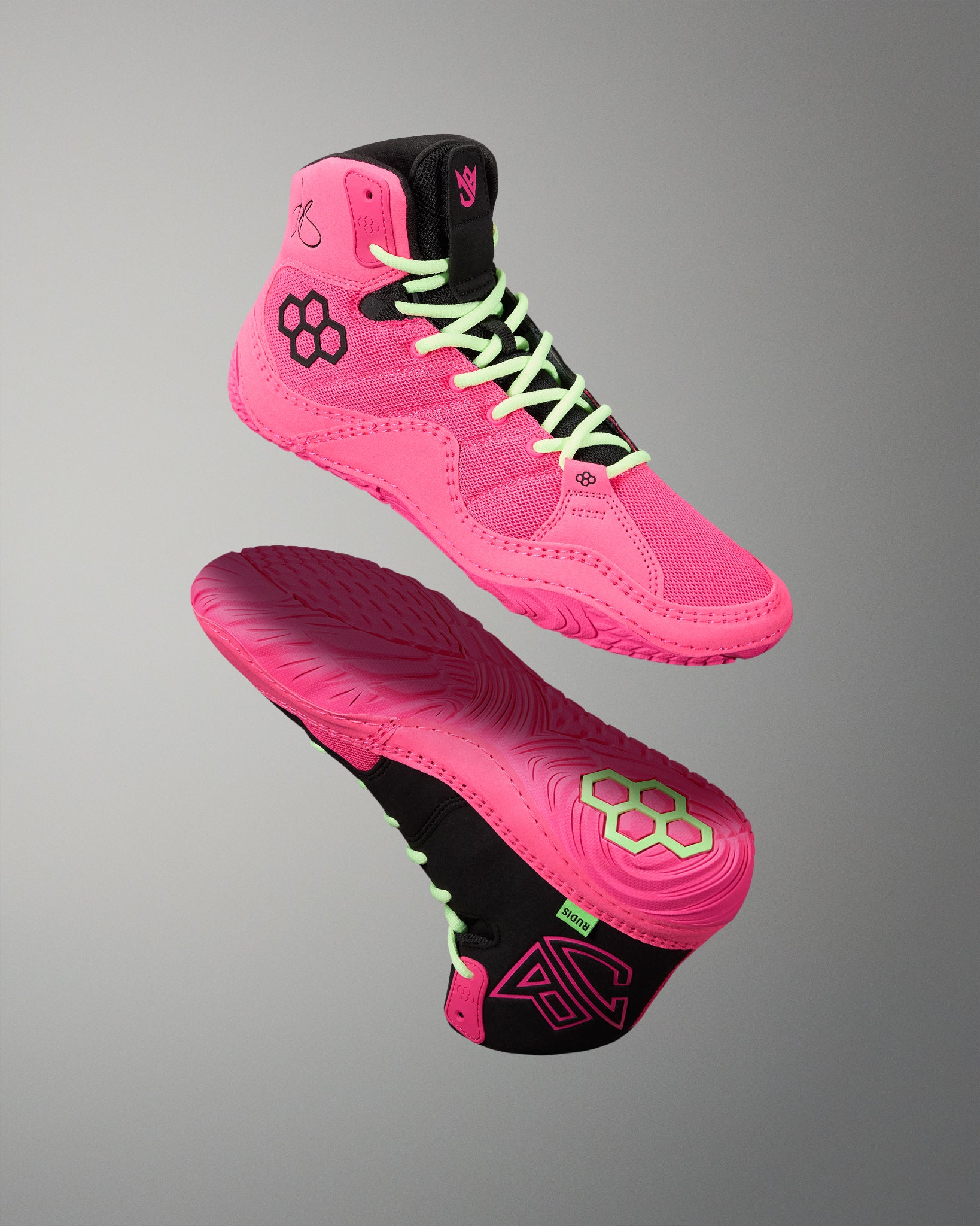 A pair of high-top athletic wrestling shoes in bright pink and black, shown floating against a neutral gray gradient background. One shoe is oriented to show the side profile with neon green laces, while the other shows the sole with a matching pink and black tread pattern.