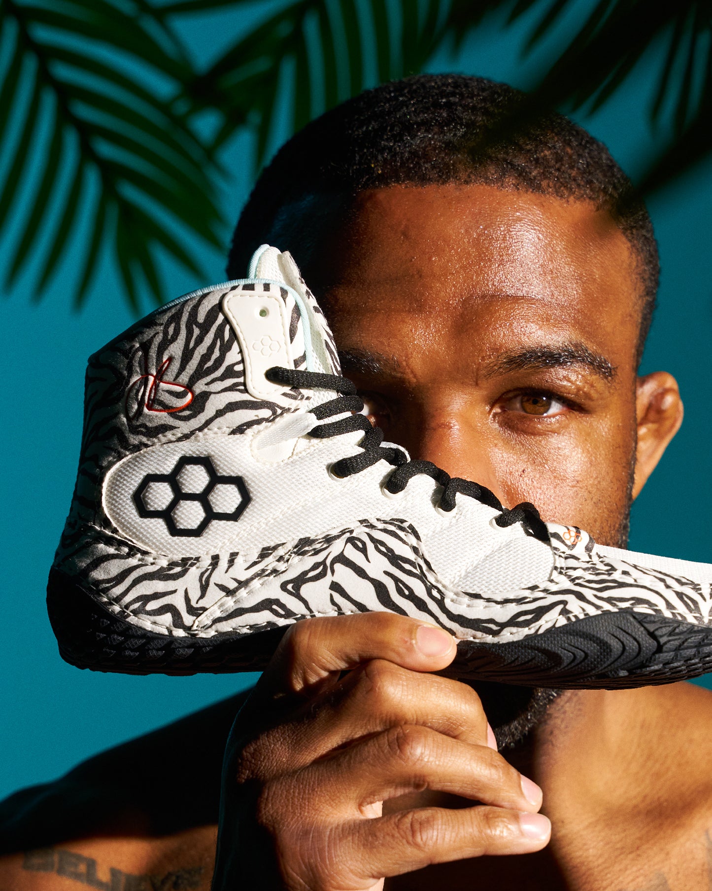 A close-up lifestyle shot of a man holding a high-top Wrestling shoe in front of his face. The wrestling shoe features a white and black zebra-stripe pattern with black laces and a black hexagonal logo on the side.