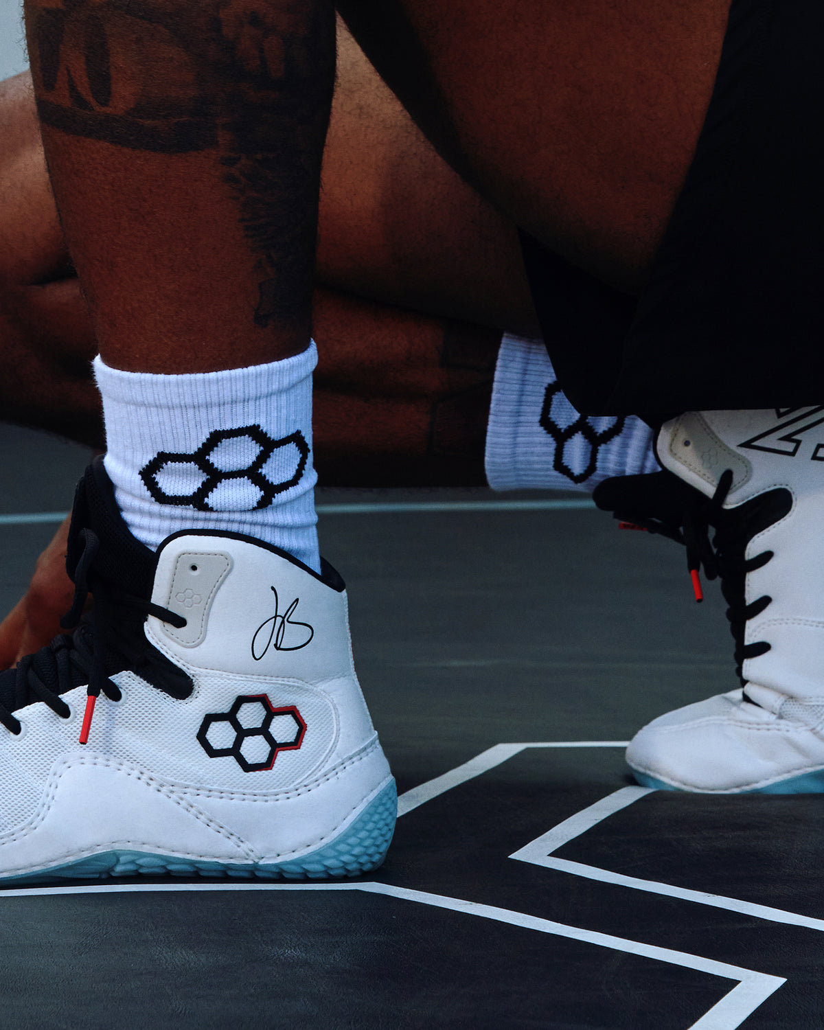 White high-top wrestling shoes with black and red geometric logo details, worn with white crew socks on a dark wrestling mat.