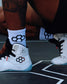 White high-top wrestling shoes with black and red geometric logo details, worn with white crew socks on a dark wrestling mat.