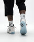 White and light blue Wrestling shoes with black graphic patterns, worn with white crew socks and black pants, shown from a rear three-quarter angle on a light gray studio background.