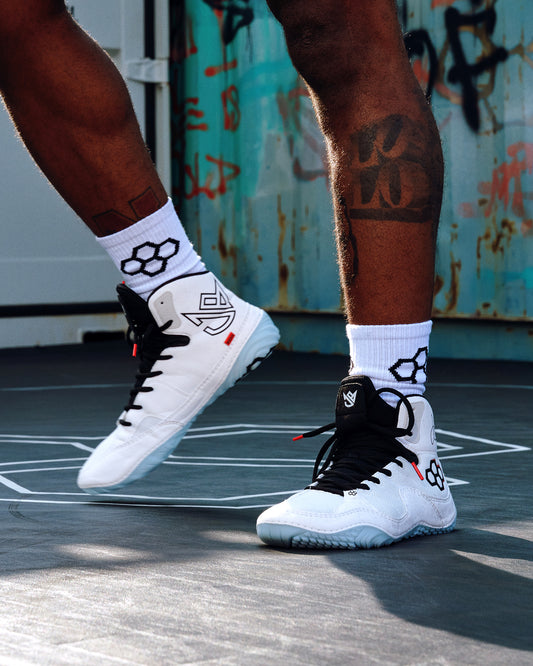 RUDIS JB1 Adult Wrestling Shoes in White Shark worn on a wrestling mat, showcasing the white high-top design, black laces, signature branding, red accents, and light blue outsole.