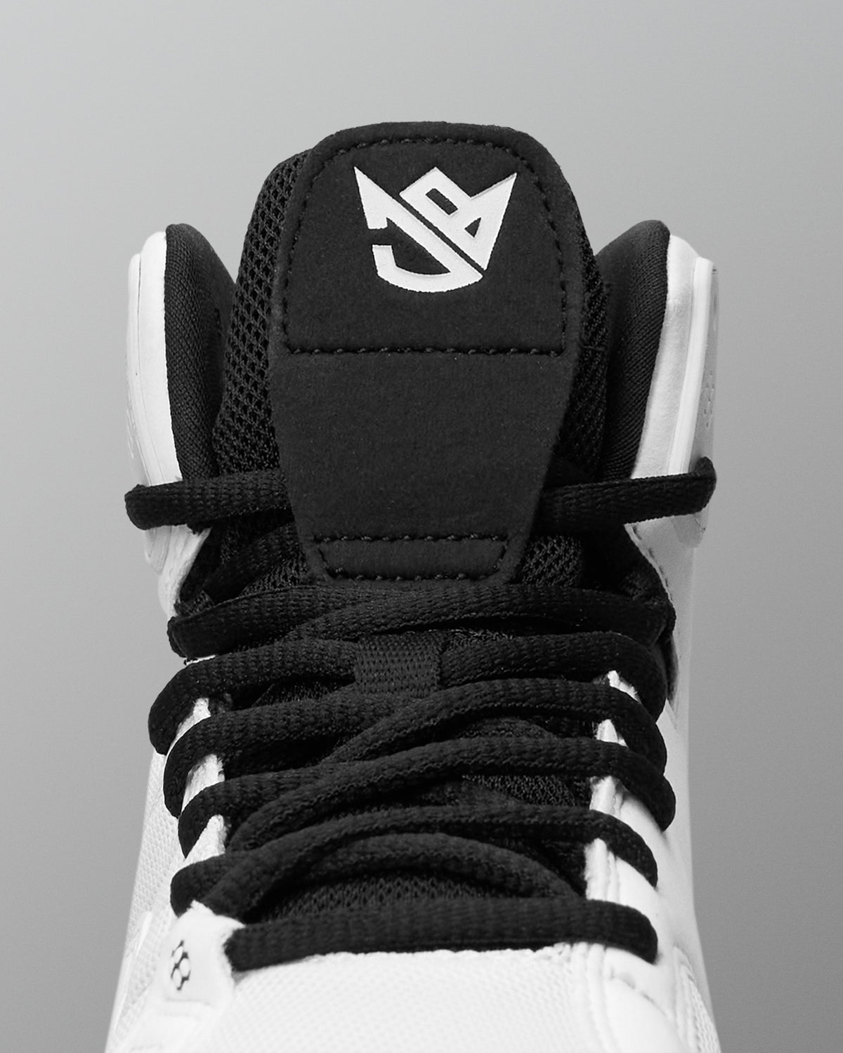 Close-up of a high-top wrestling shoes showing the black tongue patch with a white stylized logo, black laces, and white upper against a gray background.