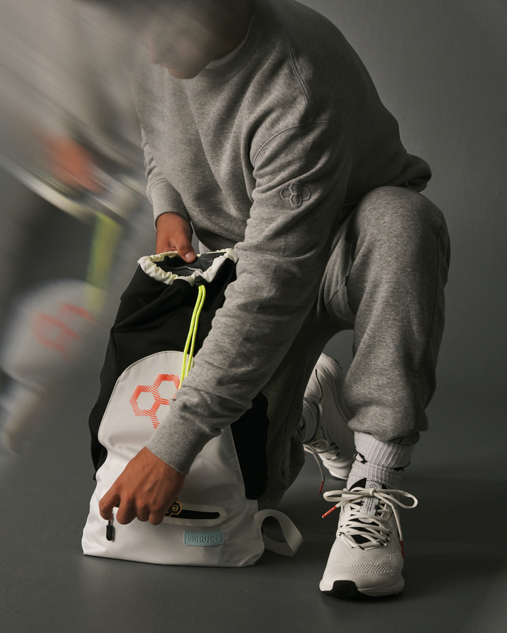 Male athlete in grey RUDIS sweatsuit and white Journey Knit Training shoes packing gear into a white and black RUDIS drawstring cinch bag with neon cord detail. 
