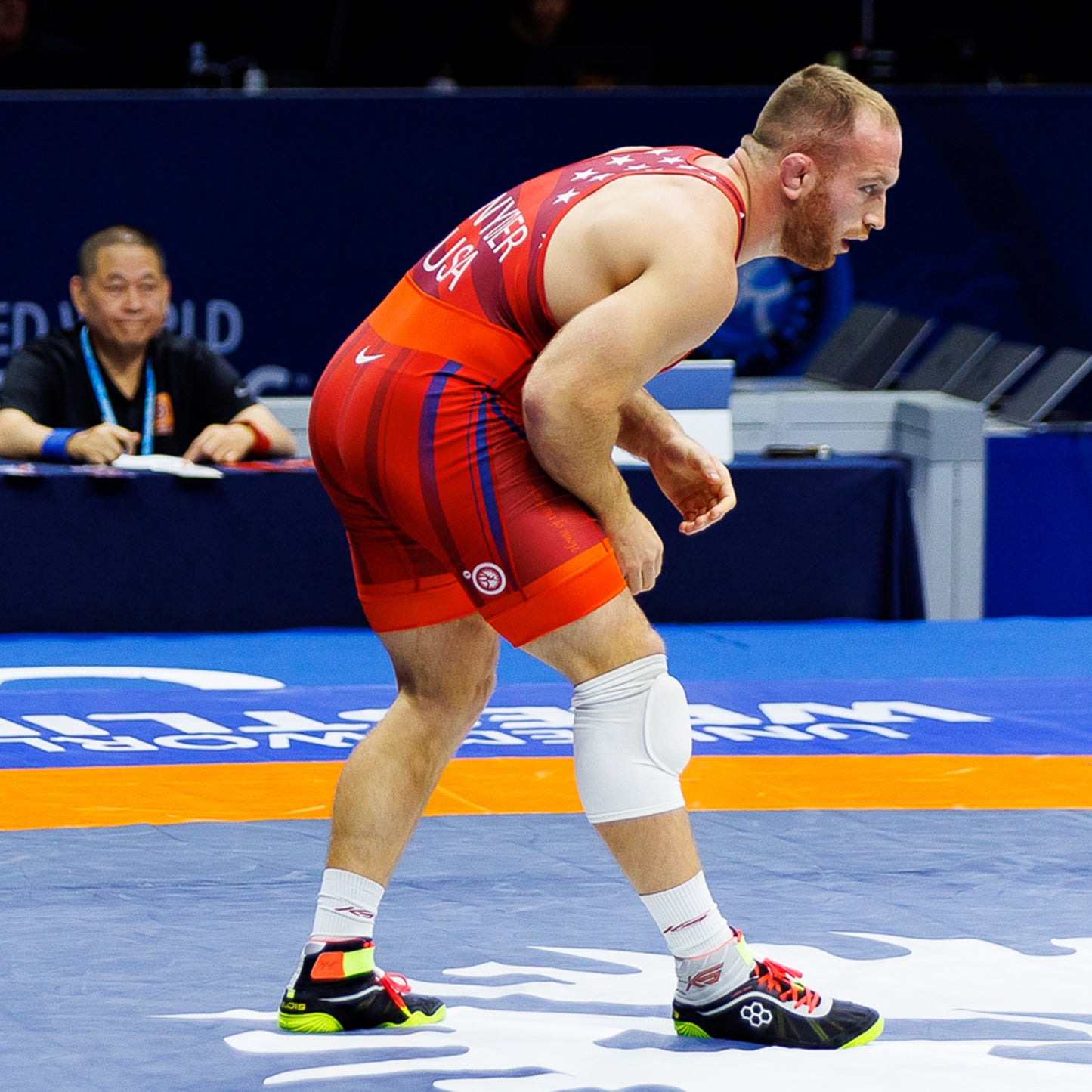 A male wrestler in a red singlet with the USA logo prepares for a match on a wrestling mat showing intense concentration and athleticism