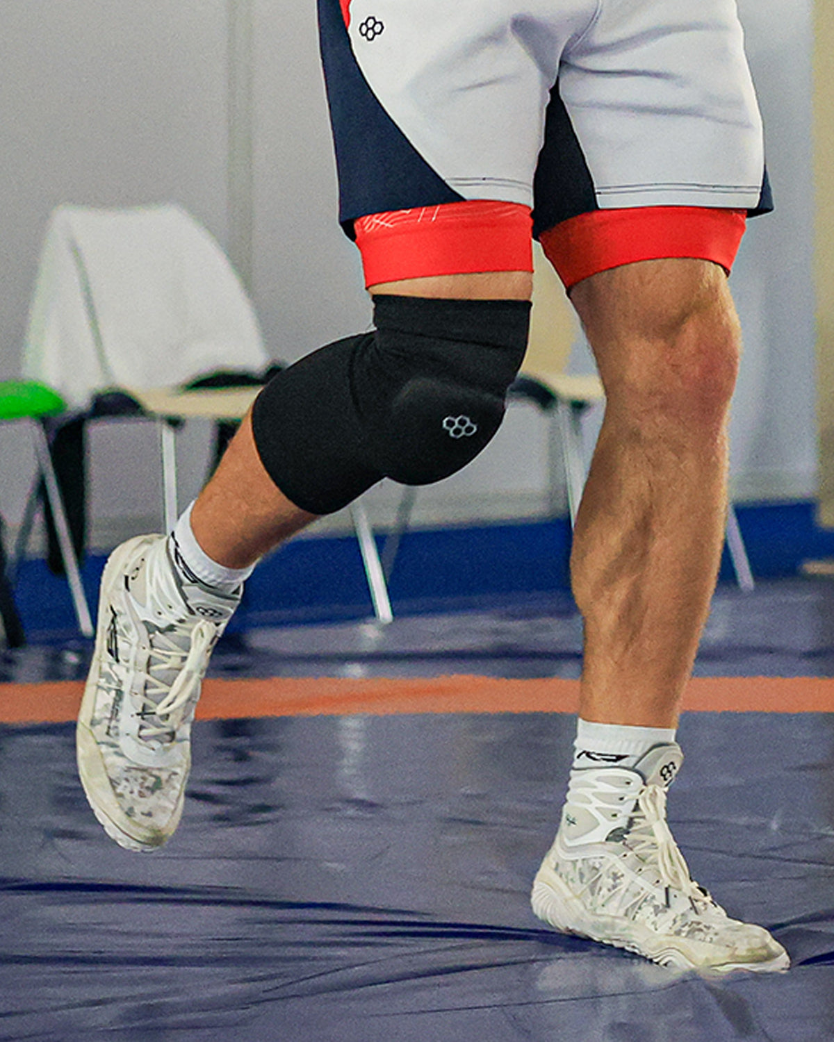 Close-up of wrestler in motion wearing black RUDIS knee pad and white RUDIS KS Infinity Edge Adult Wrestling Shoes in Winter Warfare wrestling on the mat. Image captures lower body athletic detail, highlighting knee protection and performance footwear.