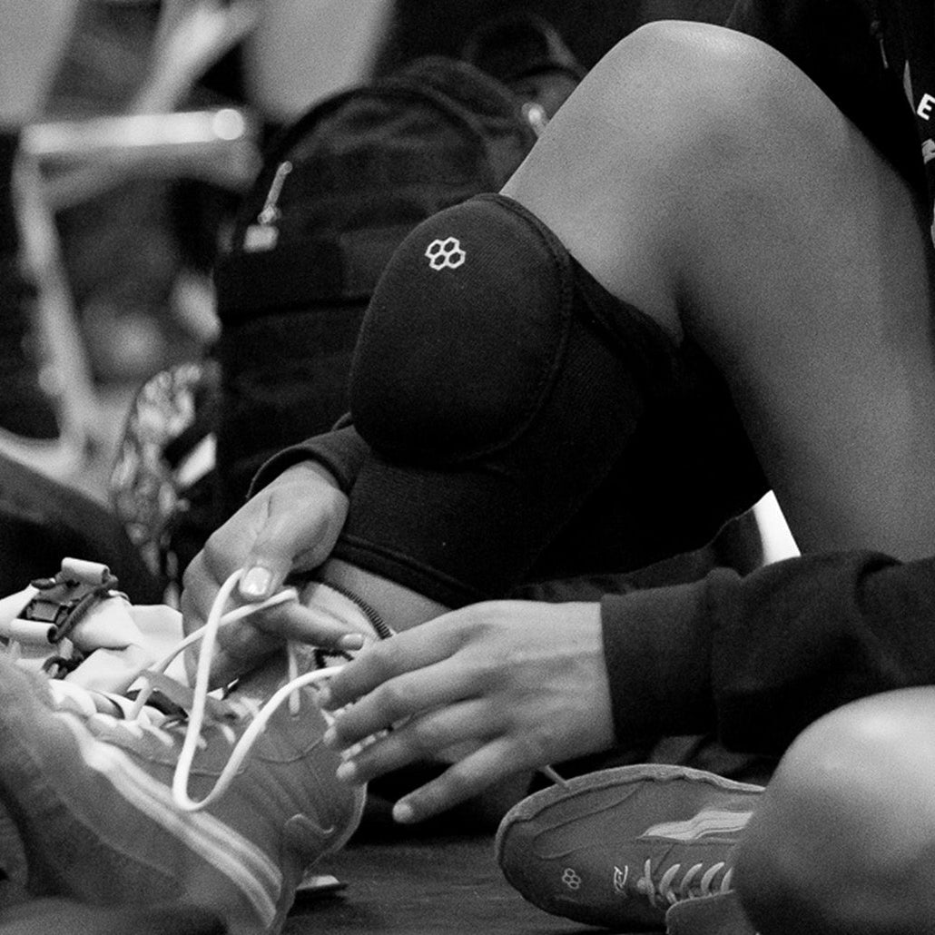 Black and white close-up of a wrestler lacing up RUDIS wrestling shoes while wearing RUDIS knee pads, capturing focus and preparation before stepping onto the mat.