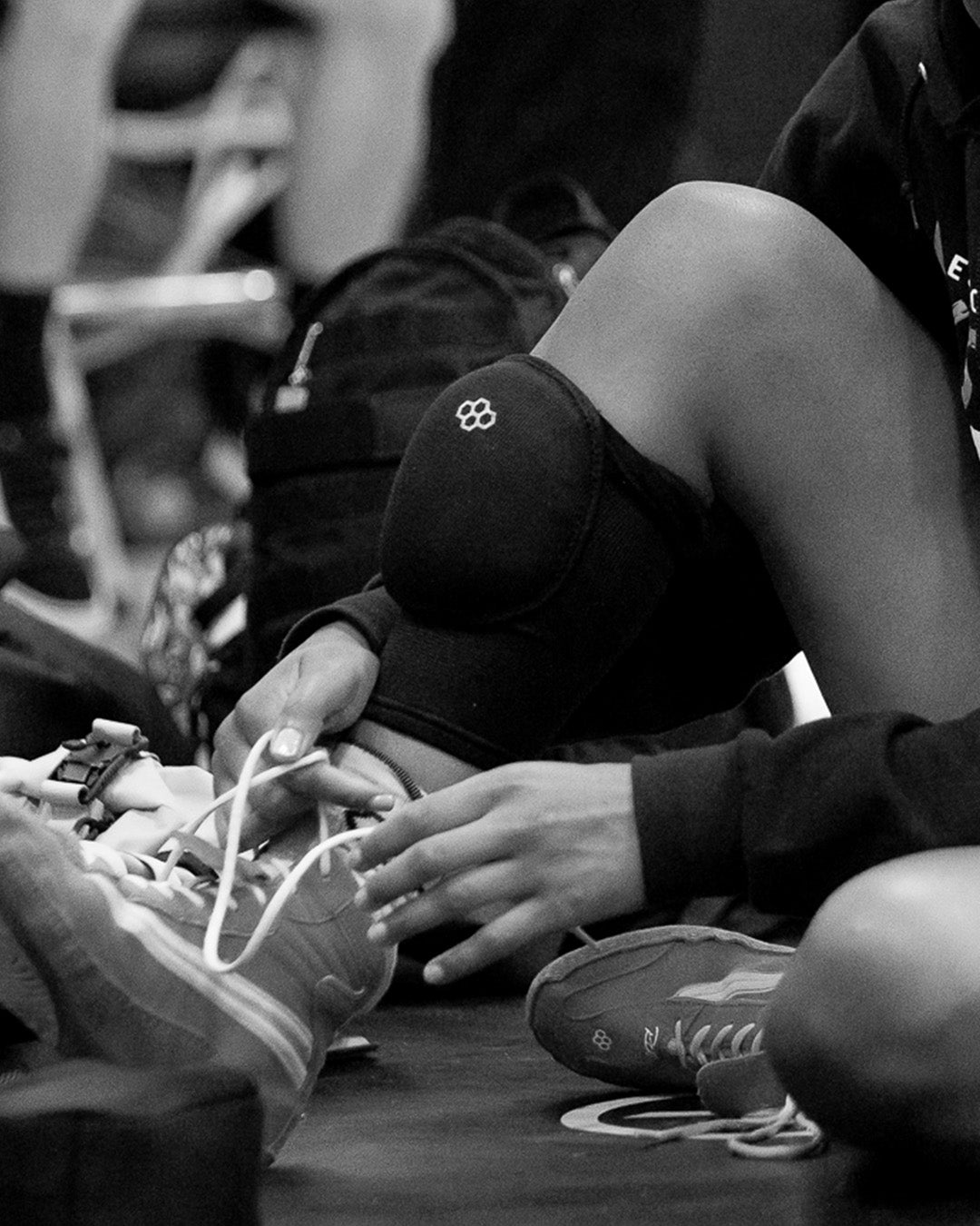 Black and white close-up of a wrestler lacing up RUDIS wrestling shoes while wearing RUDIS knee pads, capturing focus and preparation before stepping onto the mat.