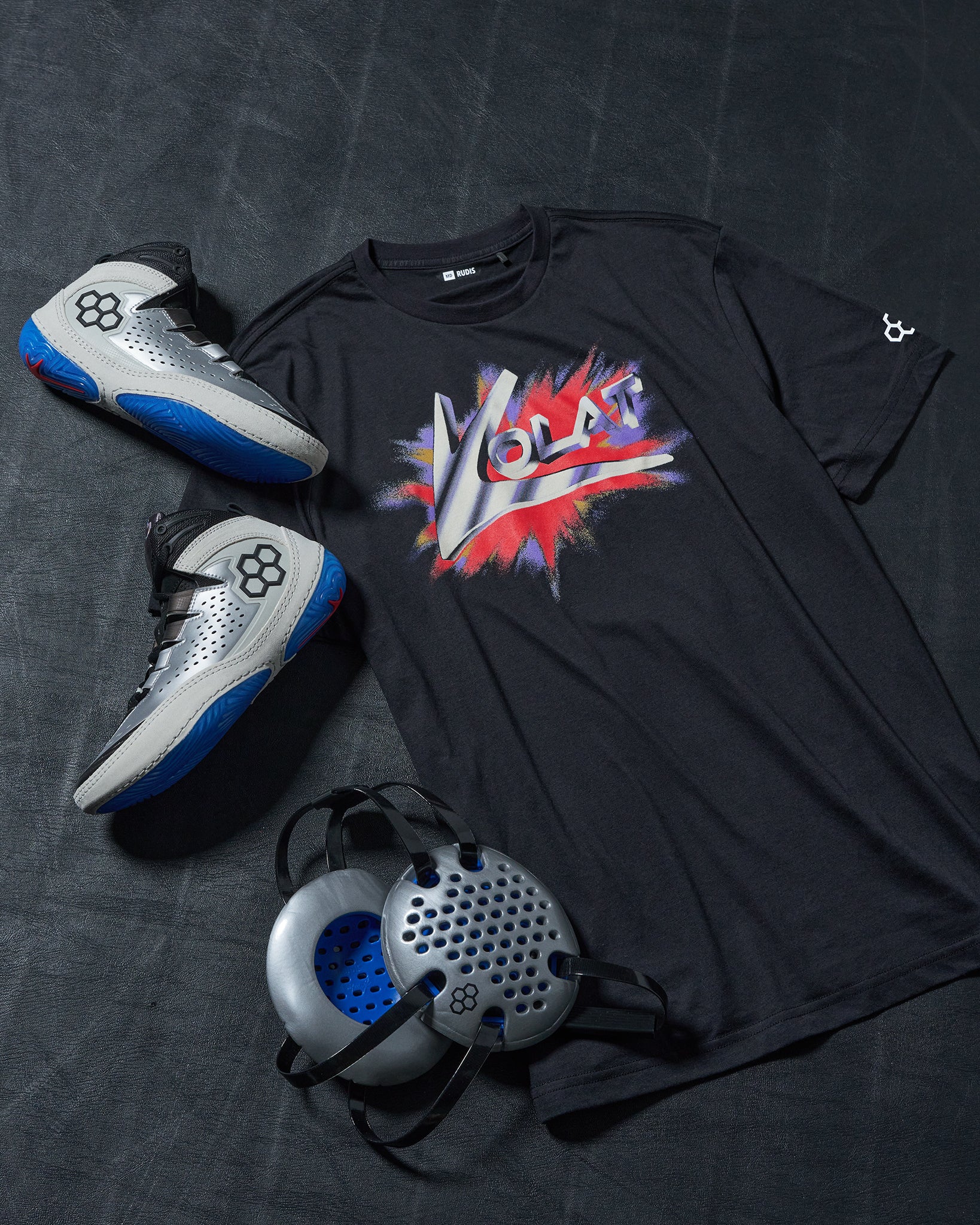 Black t-shirt with colorful graphic design, silver wrestling shoes with blue accents, and a silver headgear on a dark wrestling mat