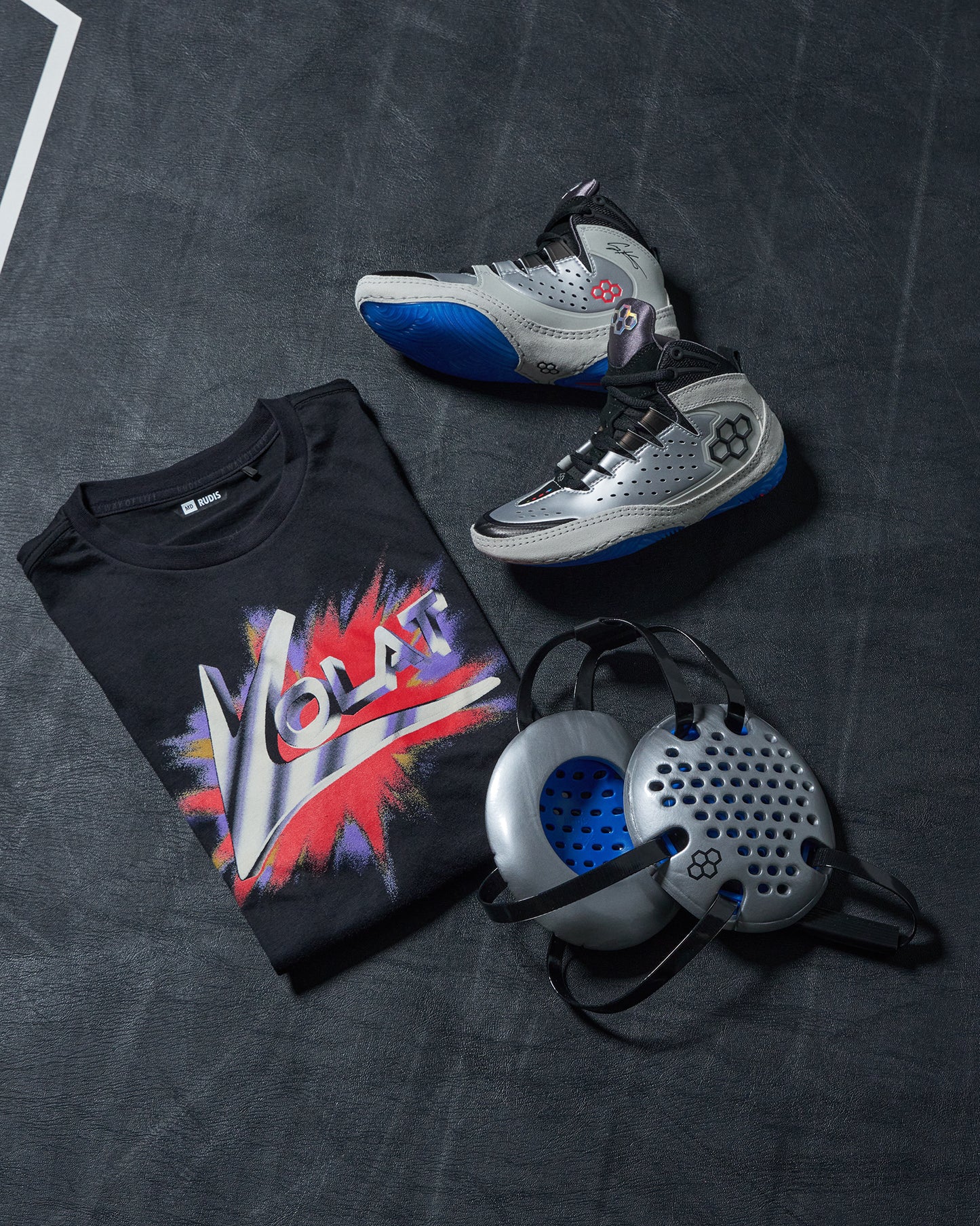 Black t-shirt with colorful graphic, silver wrestling shoes, and gray headgear on a dark wrestling mat