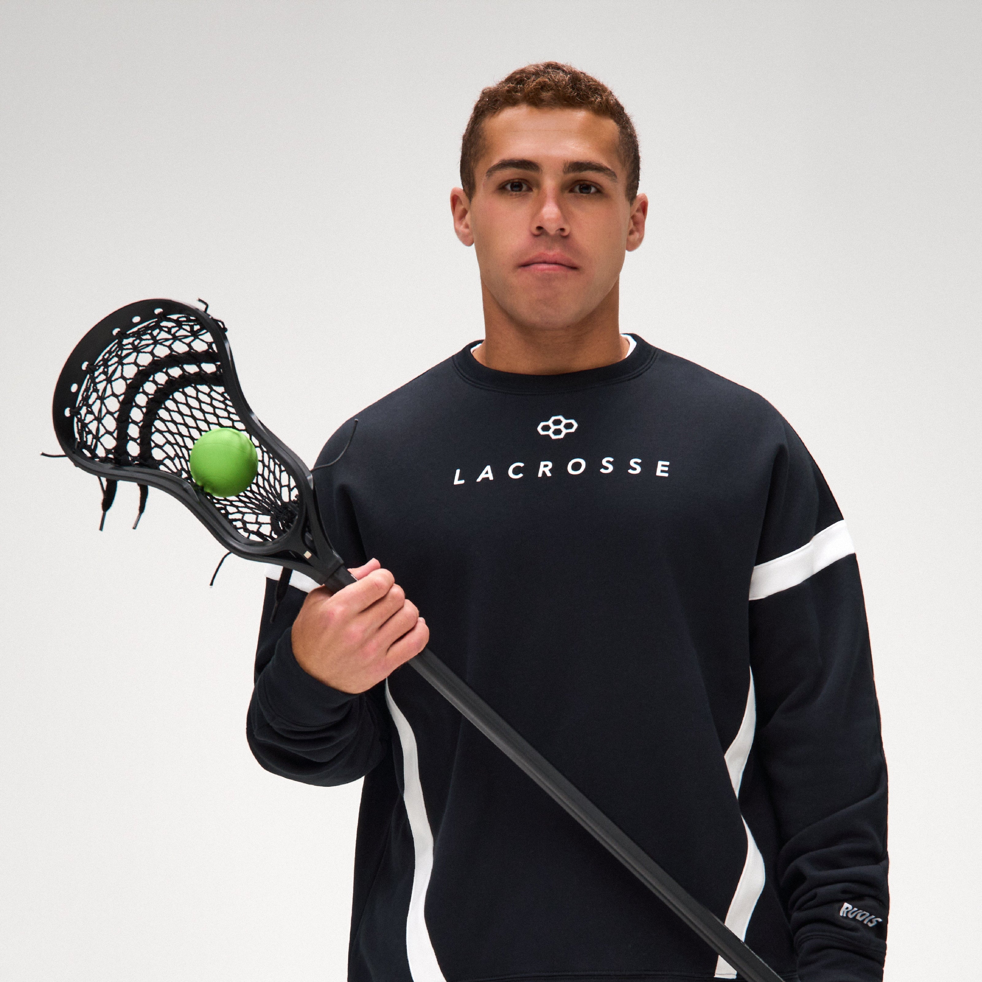 A man, Andrew McAdorey, wearing a black RUDIS Lacrosse crewneck sweatshirt with white stripes holds a lacrosse stick with a green ball, set against a plain light gray studio background.
