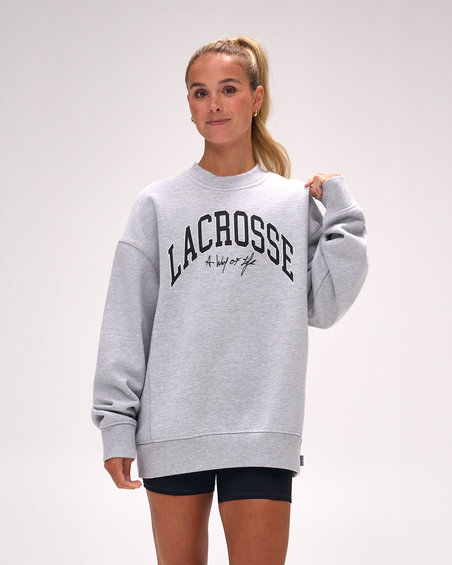 A woman wearing a light gray heather crewneck sweatshirt with 'LACROSSE' printed in black arched text across the chest, paired with black athletic shorts, against a plain light gray studio background.