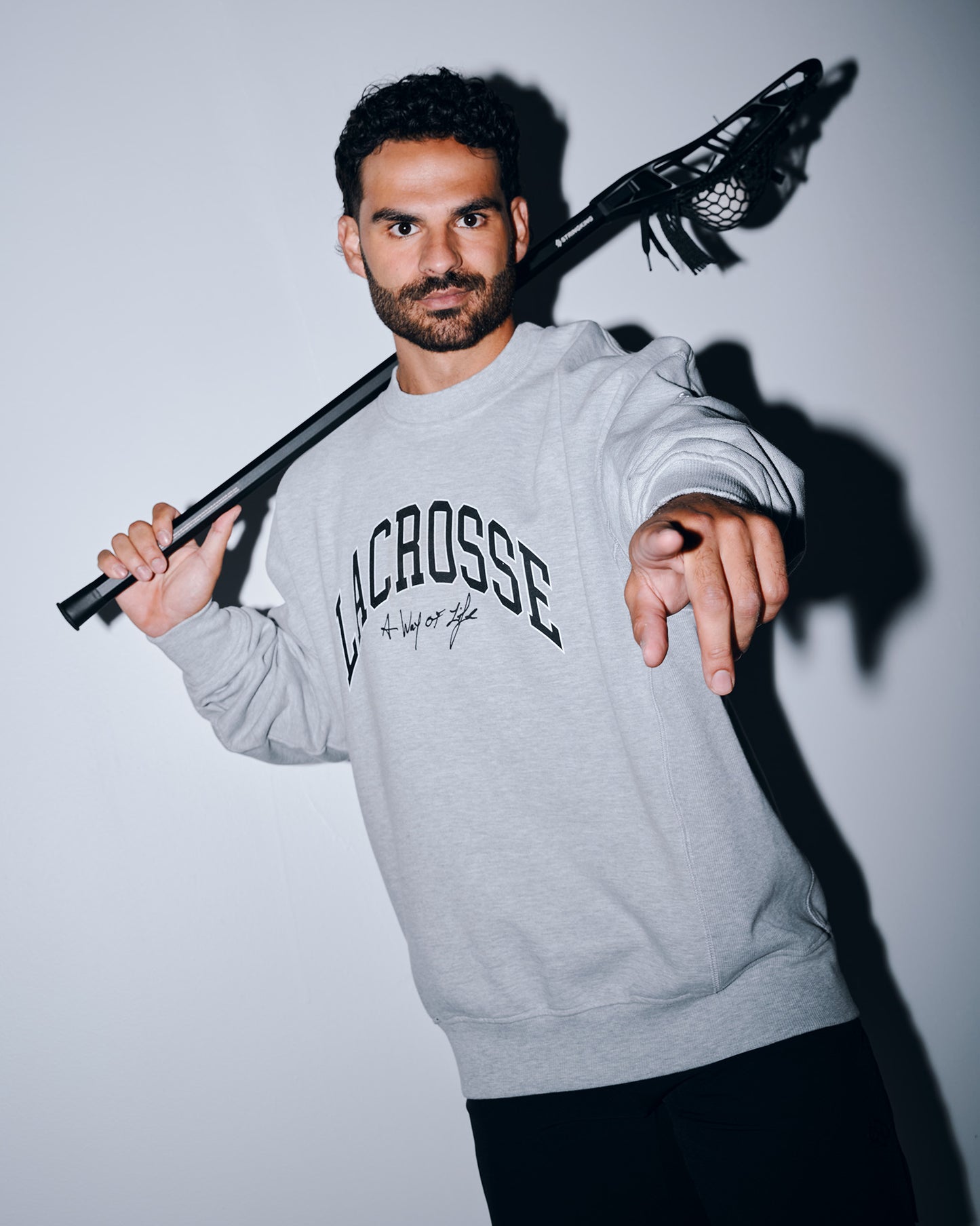 Male athlete wearing the RUDIS Motto Lacrosse Titan Heavyweight Crewneck in athletic gray while holding a lacrosse stick over his shoulder. The premium heavyweight sweatshirt features bold "Lacrosse" graphics with the signature "A Way of Life" slogan.