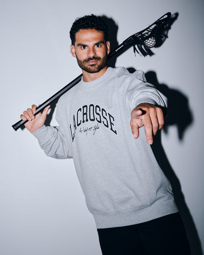 Male athlete wearing the RUDIS Motto Lacrosse Titan Heavyweight Crewneck in athletic gray while holding a lacrosse stick over his shoulder. The premium heavyweight sweatshirt features bold "Lacrosse" graphics with the signature "A Way of Life" slogan.