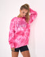 A woman wearing a bright pink tie-dye sweatshirt with 'LACROSSE' printed across the chest, paired with black shorts, posing against a plain white wall.