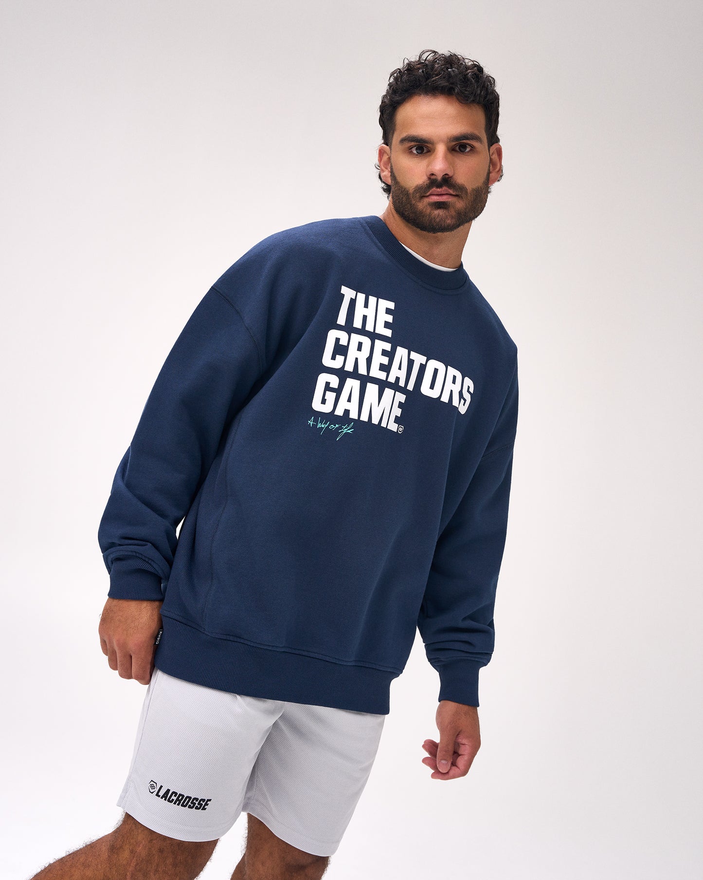 A man wearing a navy blue crewneck sweatshirt with 'THE CREATORS GAME.' printed in bold white letters on the front, paired with white shorts, photographed against a light gray studio background.