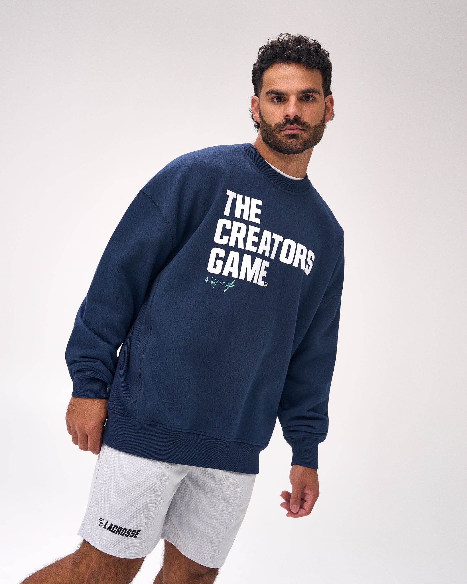 A man wearing a navy blue crewneck sweatshirt with 'THE CREATORS GAME.' printed in bold white letters on the front, paired with white shorts, photographed against a light gray studio background.
