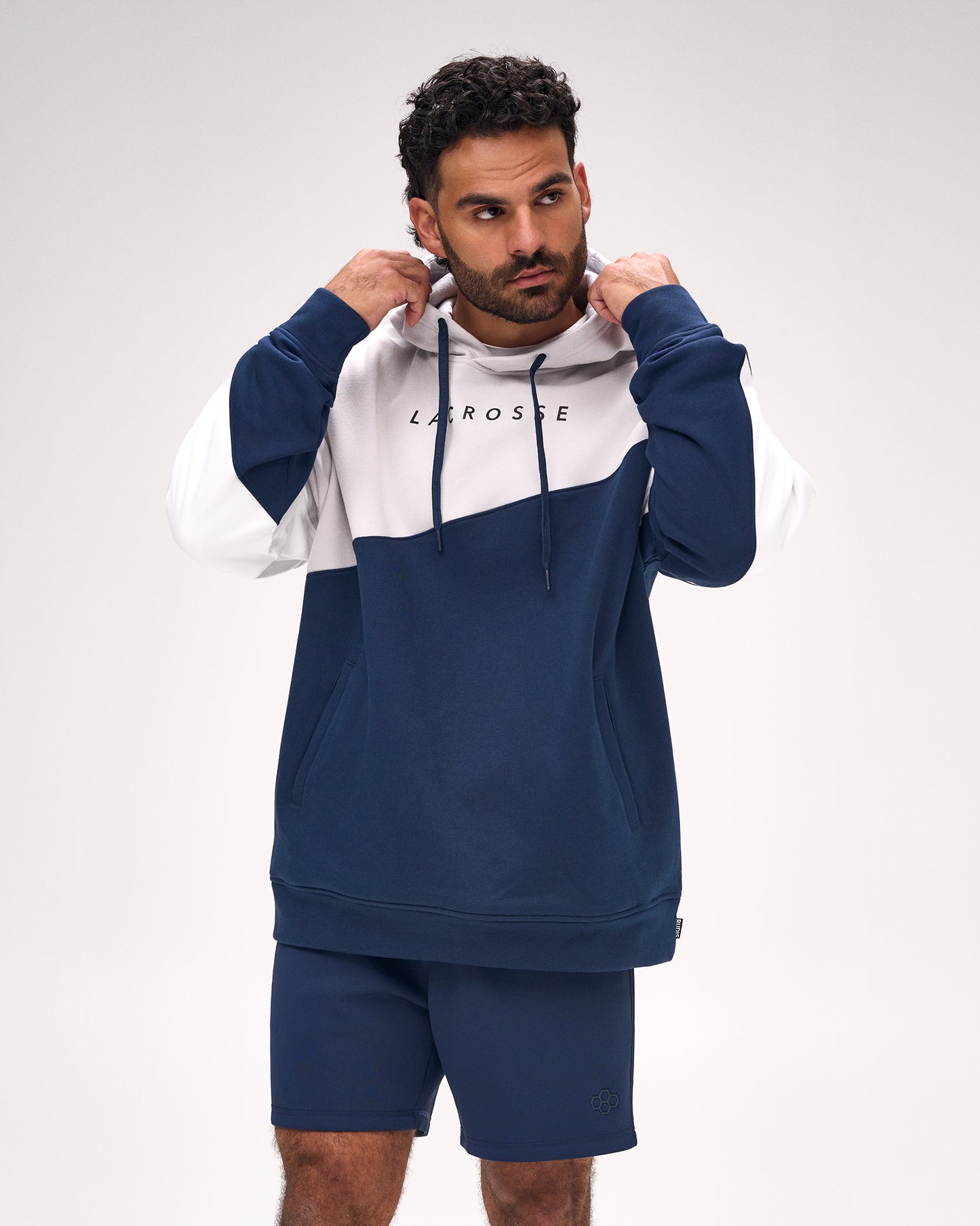 A man wearing a navy blue and white color-blocked Lacrosse hoodie with matching navy shorts, posing against a light gray studio background.