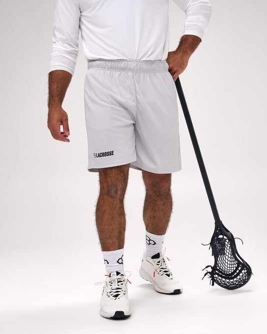 A person wearing light gray lacrosse shorts with a black 'LACROSSE' text logo on the left leg, paired with a white long-sleeve shirt, white socks, and white lacrosse shoes, holding a black lacrosse stick against a white studio background.