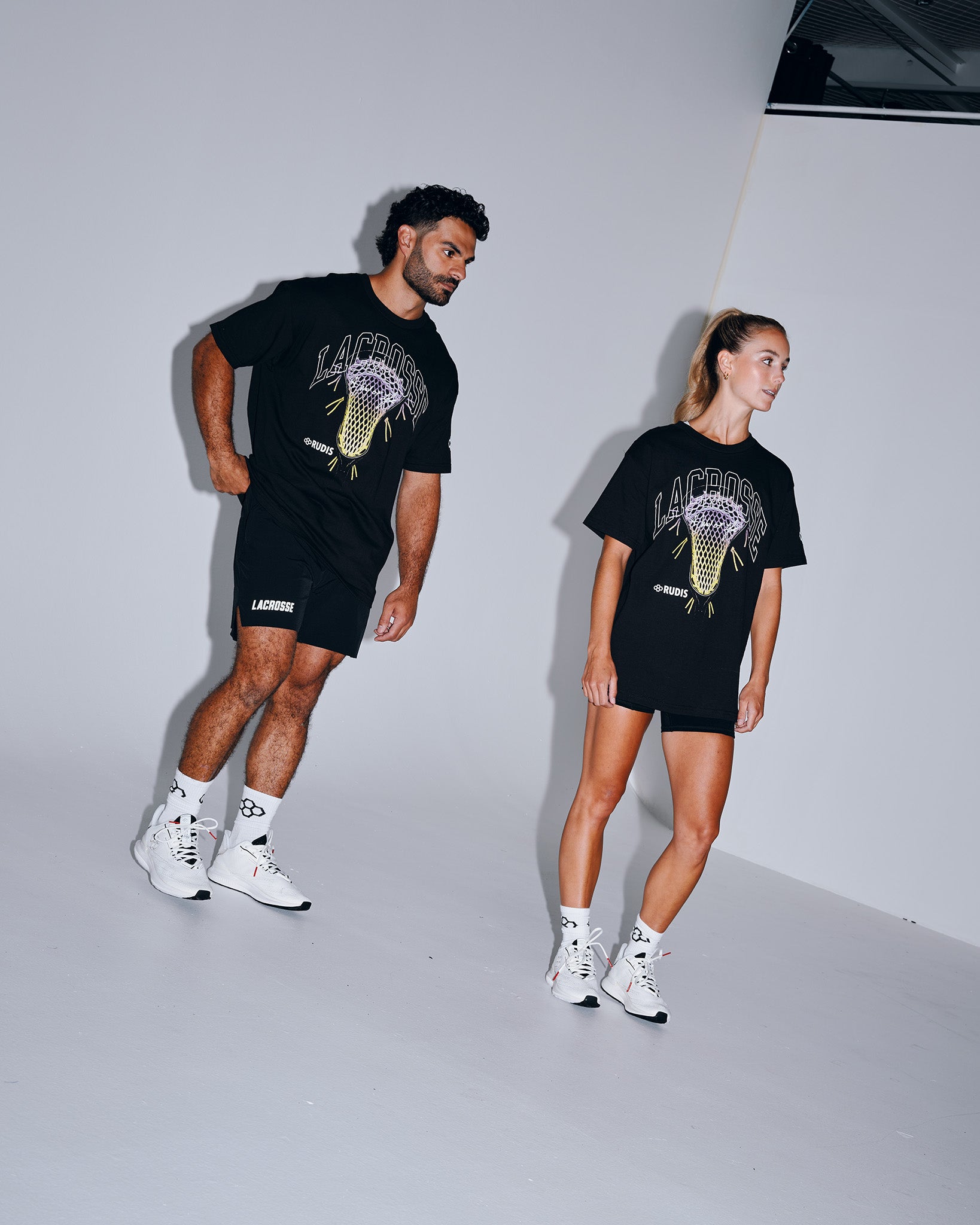 Male and female athletes wearing the RUDIS Motto Lacrosse Heavyweight T-Shirt in Black featuring a bold lacrosse head graphic, oversized fit, premium heavyweight cotton, and everyday athletic styling.