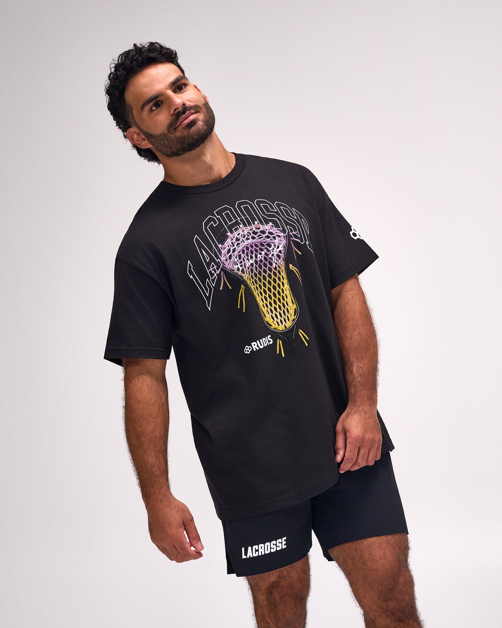 A man modeling a black Lacrosse brand t-shirt with a yellow and purple lacrosse graphic, paired with black shorts, against a light gray studio background.