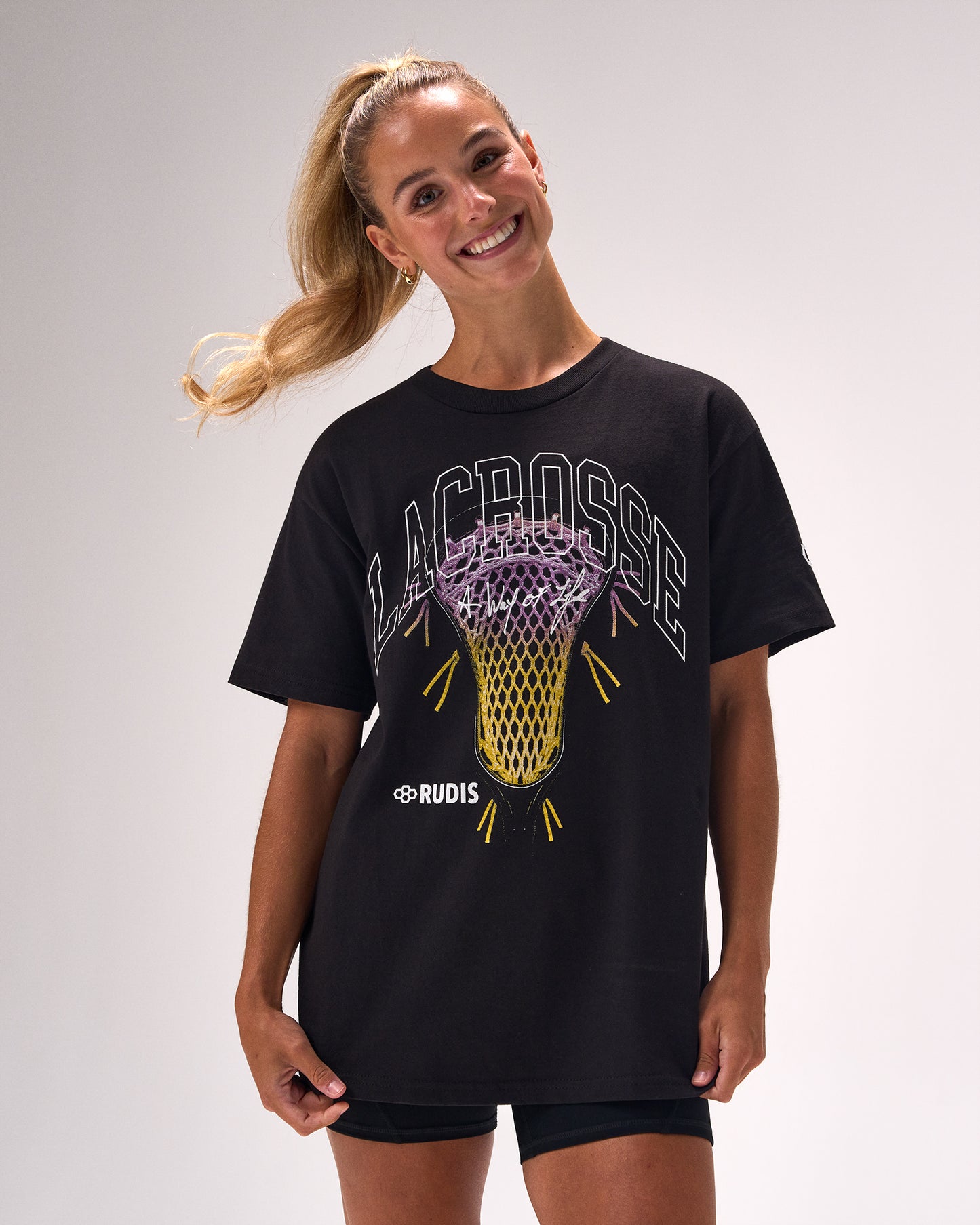 A woman modeling a black short-sleeve t-shirt with a graphic of a lacrosse net and the word 'LACROSSE' in white and purple, with 'RUDIS' printed below it, against a light gray studio background.
