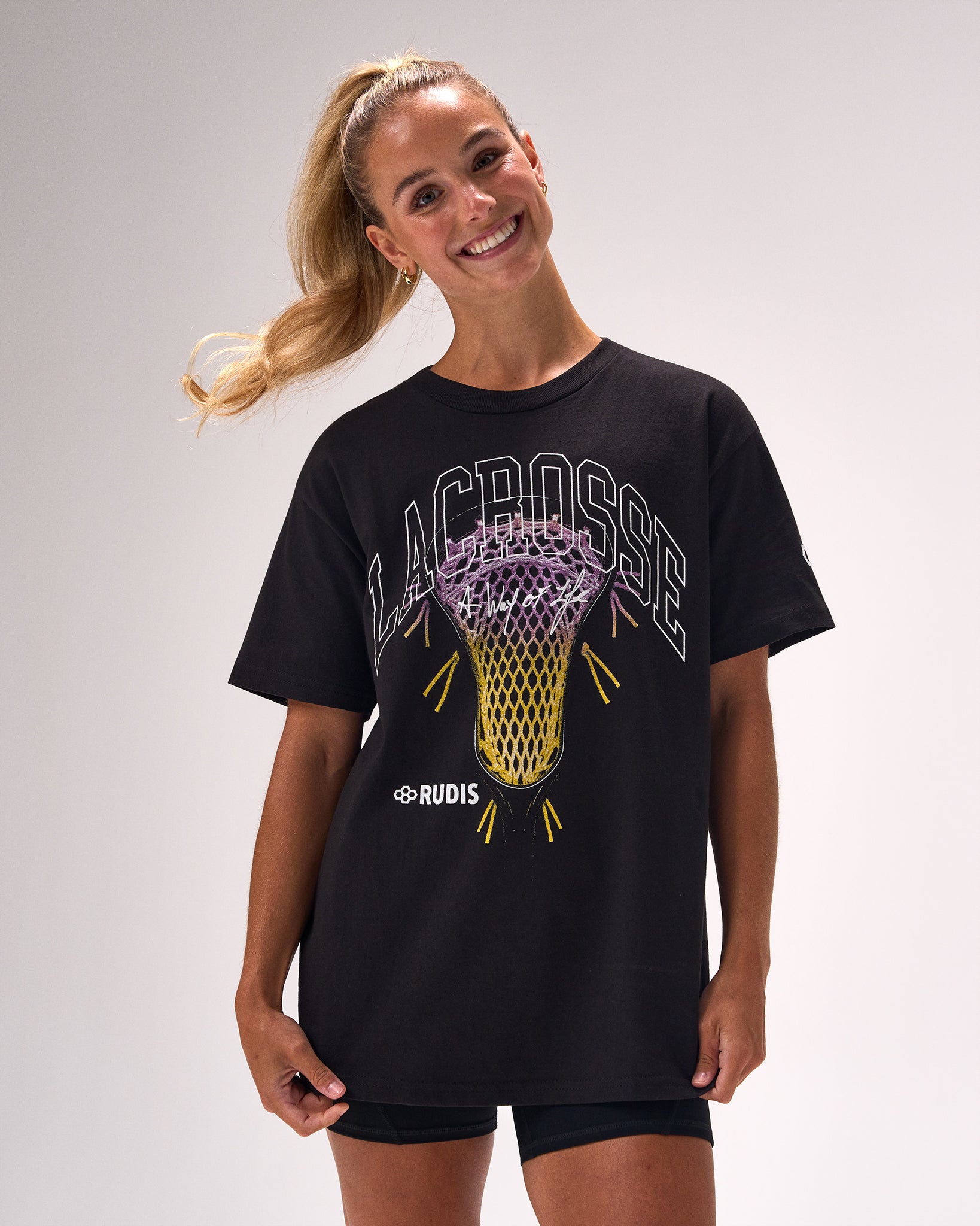 A woman modeling a black short-sleeve t-shirt with a graphic of a lacrosse net and the word 'LACROSSE' in white and purple, with 'RUDIS' printed below it, against a light gray studio background.