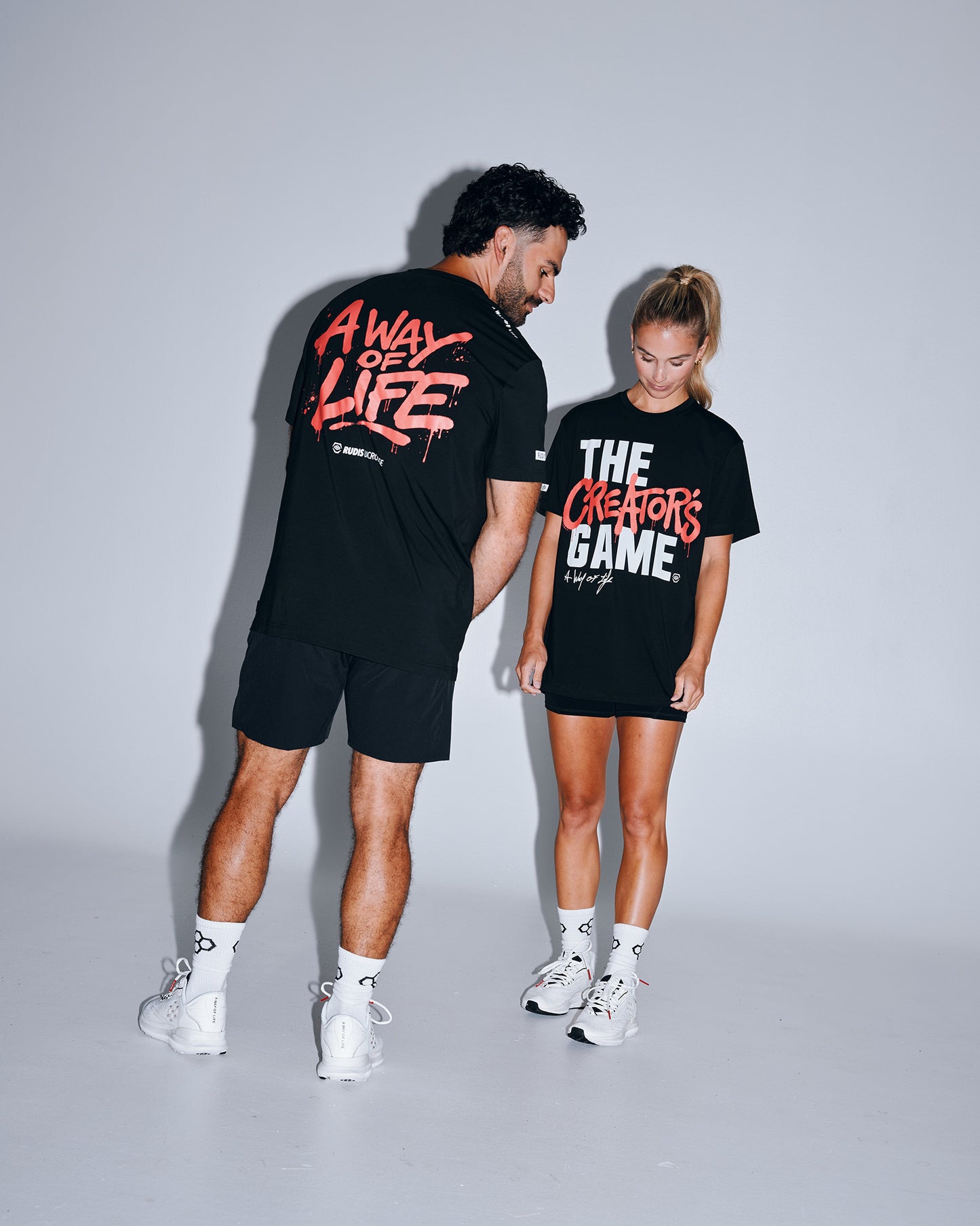 Male and female athletes wearing the RUDIS The Creator's Game Hybrid T-Shirt in black. The front features bold "The Creator's Game" artwork, while the back showcases oversized "A Way of Life" graphics inspired by lacrosse culture.
