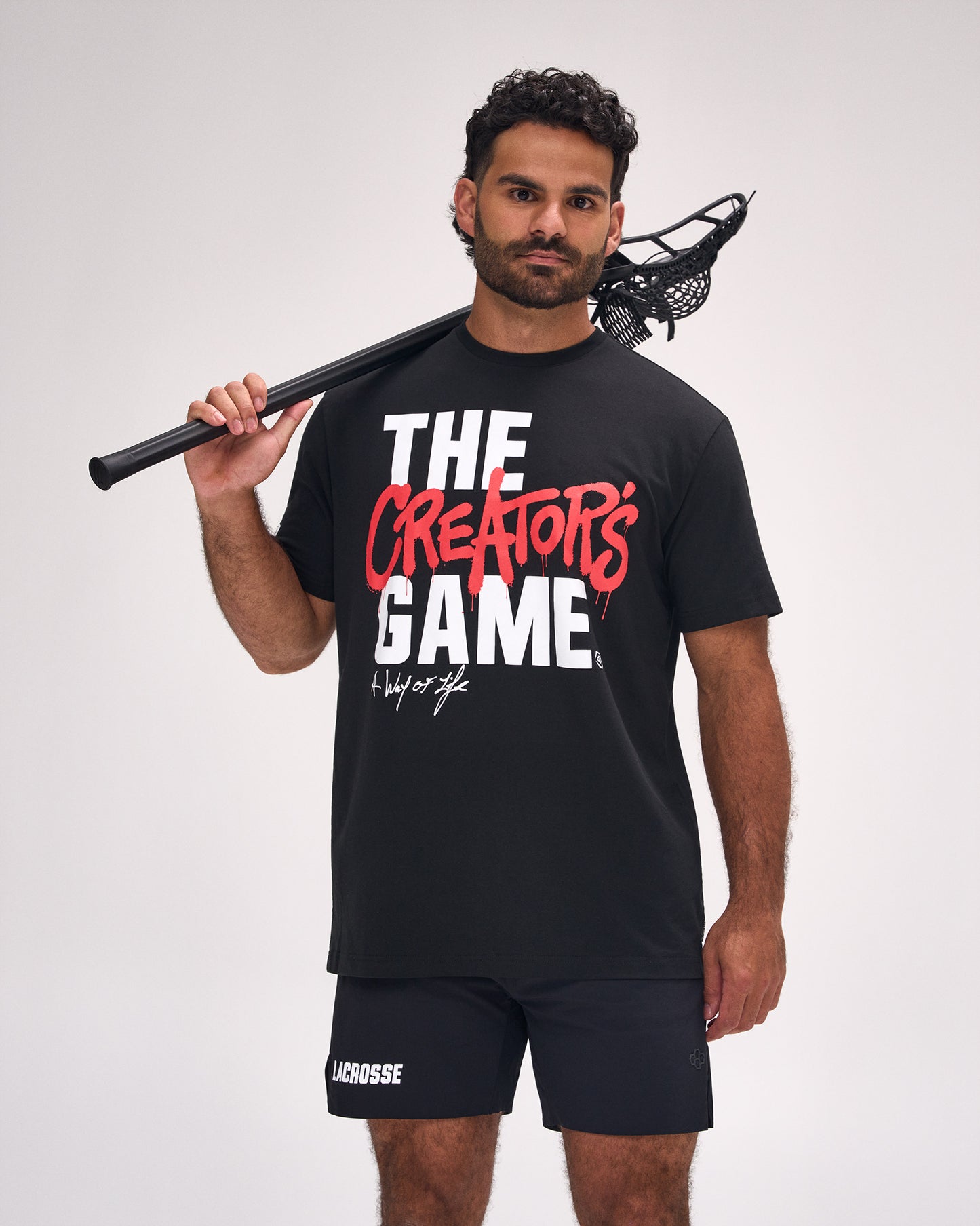 Male athlete wearing the RUDIS The Creator's Game Hybrid T-Shirt in Black featuring bold "The Creator's Game" graphic and relaxed everyday fit while holding a lacrosse stick.