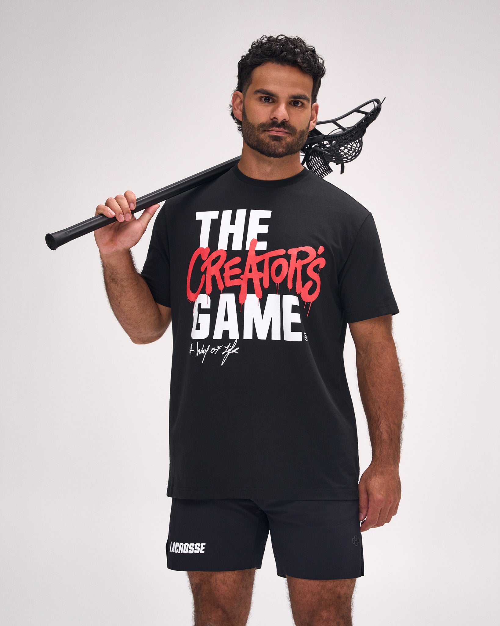 Male athlete wearing the RUDIS The Creator's Game Hybrid T-Shirt in Black featuring bold "The Creator's Game" graphic and relaxed everyday fit while holding a lacrosse stick.