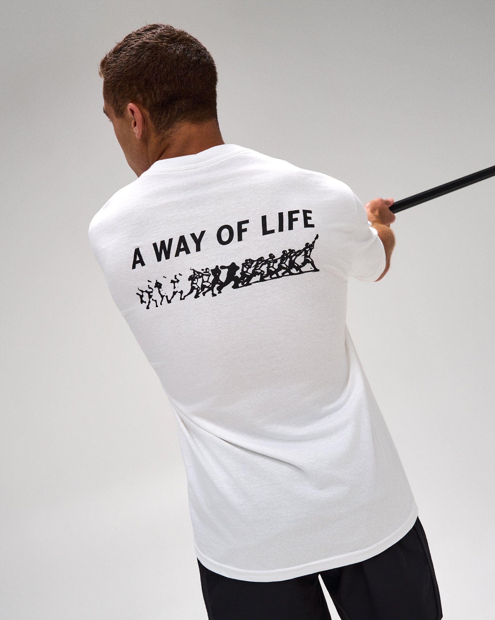 A man viewed from behind wearing a white t-shirt with a black graphic print that reads 'A WAY OF LIFE' and shows a black staff or rod extending to the right against a light gray studio background.