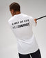 A man viewed from behind wearing a white t-shirt with a black graphic print that reads 'A WAY OF LIFE' and shows a black staff or rod extending to the right against a light gray studio background.