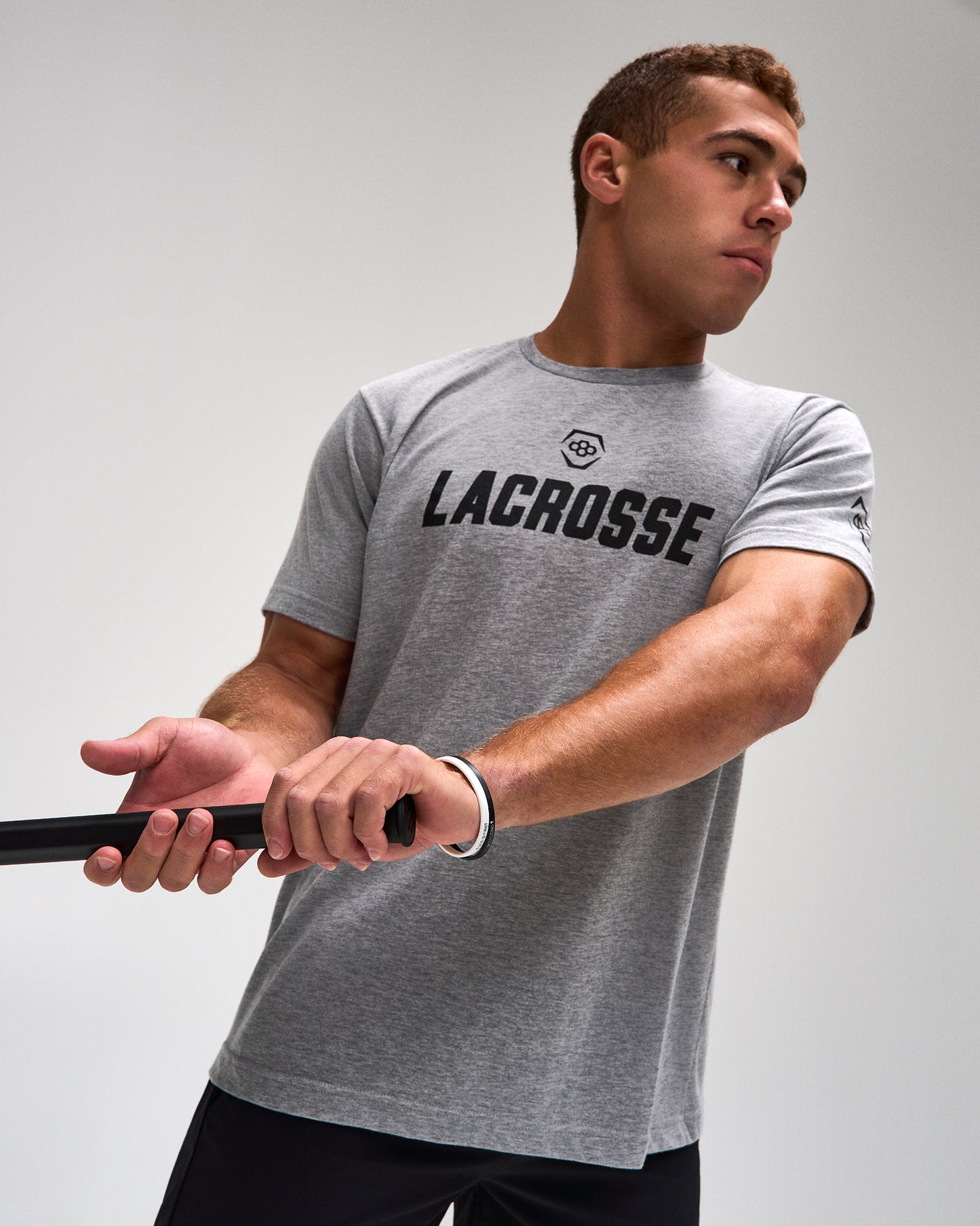 A man wearing a heather gray athletic t-shirt with 'LACROSSE' printed in black across the chest, holding a lacrosse stick against a light gray studio background.