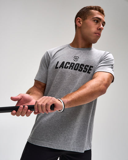 A man wearing a heather gray athletic t-shirt with 'LACROSSE' printed in black across the chest, holding a lacrosse stick against a light gray studio background.