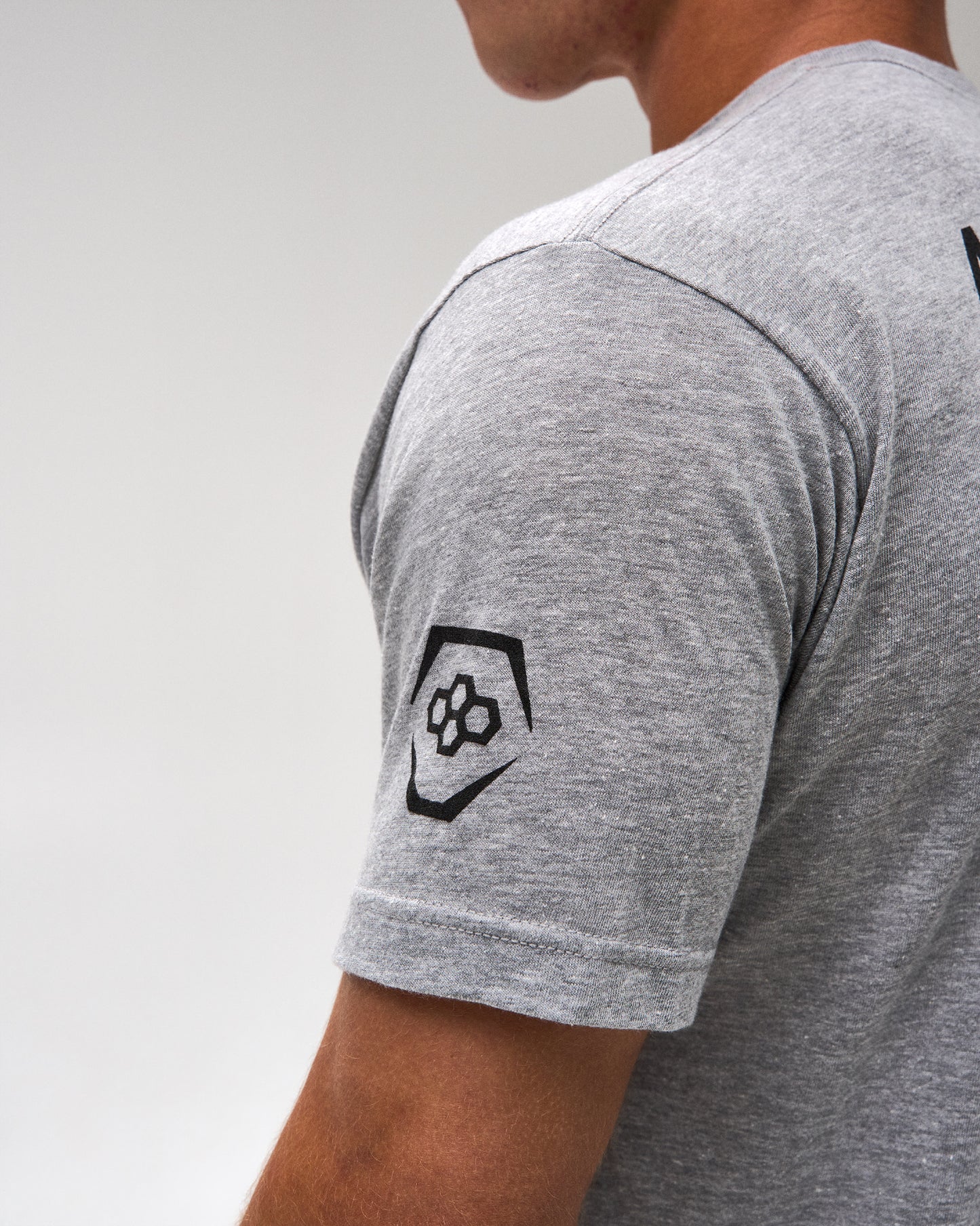 Close-up of a person wearing a heather gray short-sleeve t-shirt, showing a black geometric logo patch on the left sleeve against a light gray background.