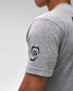 Close-up of a person wearing a heather gray short-sleeve t-shirt, showing a black geometric logo patch on the left sleeve against a light gray background.