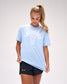 Female athlete wearing the RUDIS LAX Motto Elite Super Soft T-Shirt in Light Blue featuring arched "Lacrosse" lettering, lacrosse head graphic, and signature "A Way of Life" artwork.